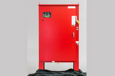 A red industrial control panel box with a digital display and multiple labels on its door, standing on a black fabric.