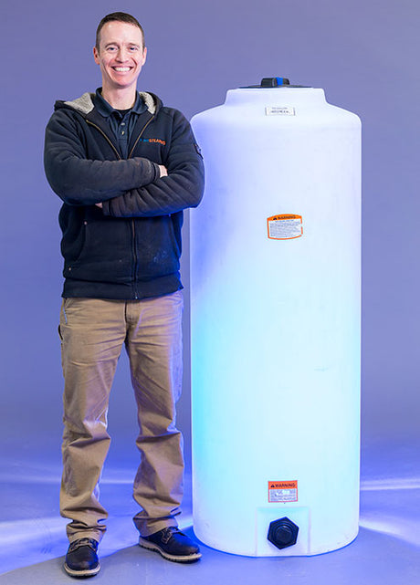 A person stands next to a Norwesco Vertical 105 Gallon storage tank on a blue-lit background. Dressed in a black jacket, brown pants, and blue shoes, they smile with arms crossed. The polyethylene resin tank features labels and a black valve at the base.