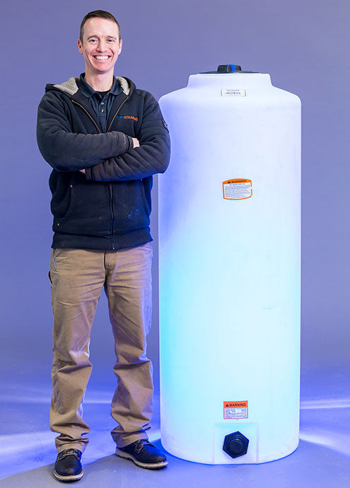 A person stands next to a Norwesco Vertical 105 Gallon storage tank on a blue-lit background. Dressed in a black jacket, brown pants, and blue shoes, they smile with arms crossed. The polyethylene resin tank features labels and a black valve at the base.