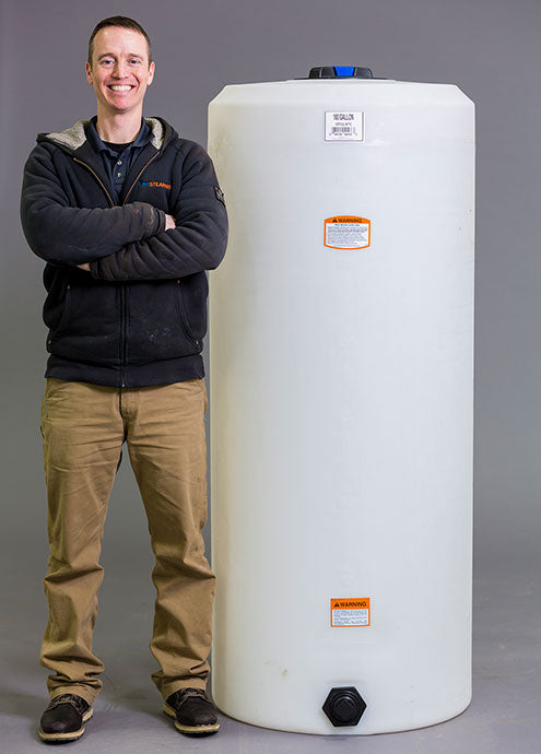 A person stands smiling with arms crossed next to a Vertical 160 Gallon liquid storage tank by Norwesco. The durable cylindrical tank features warning labels and a black cap on top. The individual, dressed in a black jacket and khaki pants, poses against a gray background.