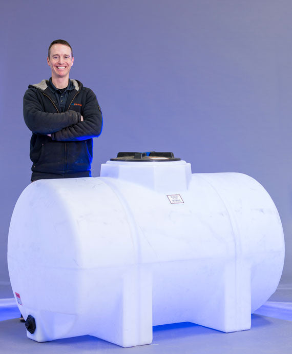 A person stands smiling with arms crossed next to a large white horizontal HORIZ. LEG 225 Gallon tank by Norwesco, set against a gray background. The cylindrical tank, resembling a leg tank, features a flat top with a hatch or cap.