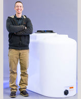 Beside a large Freestanding Doorway 300 Gallon tank by Norwesco, a man stands confidently with his arms crossed, smiling. His attire includes a dark jacket, khaki pants, and black shoes. The tank features a black cap and bears its label at the bottom right corner, standing out against the gray background that complements his assured presence.