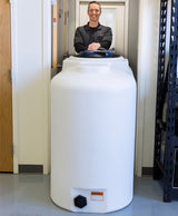 A person stands behind the Freestanding Doorway 300 Gallon, a large white cylindrical water tank from Norwesco with a blue lid, seamlessly blending into the room's minimalist aesthetic. The space is characterized by white walls and a gray floor, with a black metal shelf positioned to the right near the door.