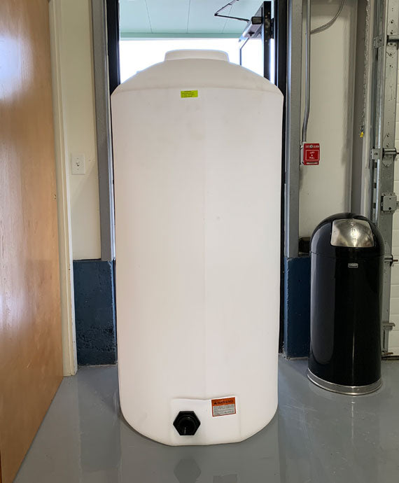 In a room with a shiny floor, the Freestanding Doorway 500 Gallon by Norwesco, a large white cylindrical storage tank, stands alongside a trash can near a partially open garage door. A small black nozzle at the bottom and its label are distinct features. Remarkably, the design suggests it could easily be adaptable to various shaped tank configurations.