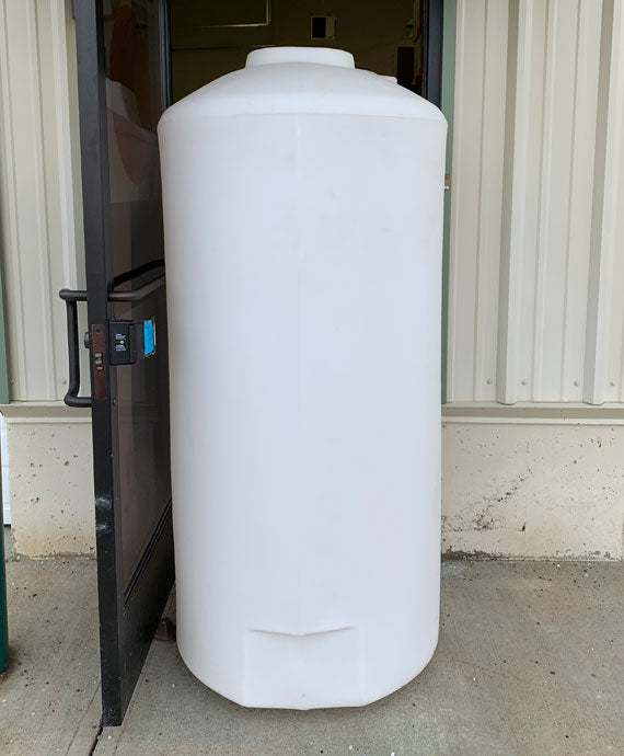 A Freestanding Doorway 500 Gallon tank from Norwesco, large, white, and rectangular in shape, is positioned vertically and partially obstructs a glass doorway entrance of a building with light-colored siding. The tank stands on a concrete surface, awkwardly fitting into the doorway space.